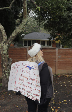 Breimer stands outside of Rice’s house, wearing her sign in protest of traffic signals, “highway-sized poles,” “arms” on poles, oversized long street signals, and “no fair open progress.”
