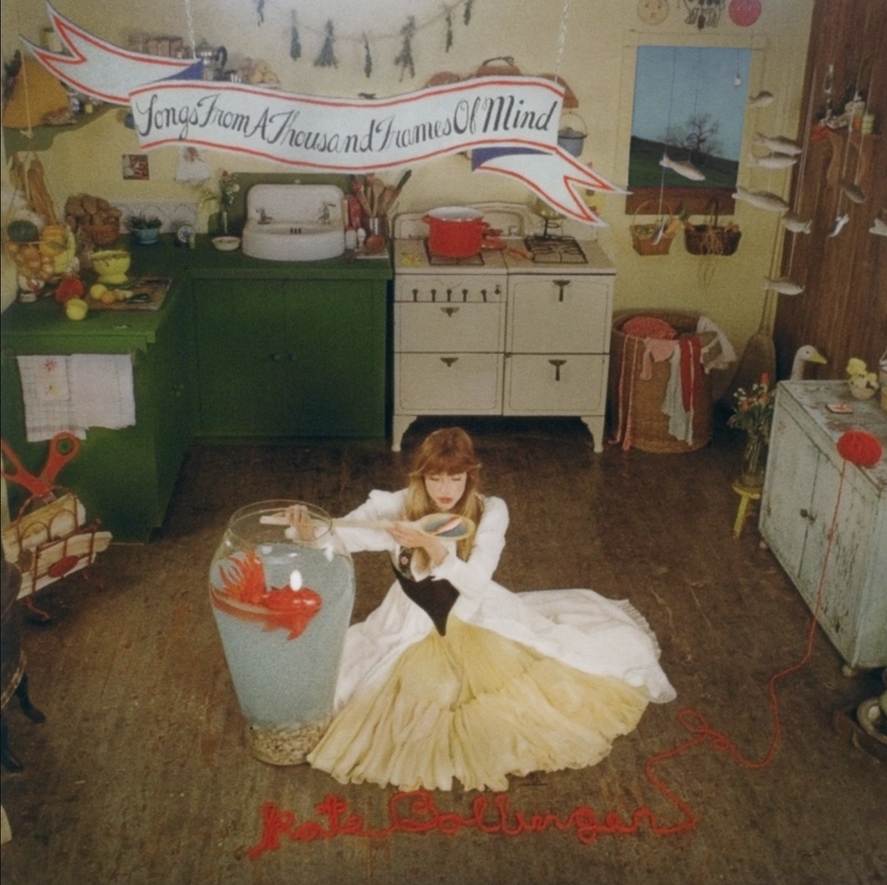 The cover for Kate Bollinger’s new album, Songs From A Thousand Frames Of Mind, depicts her sitting on the kitchen floor surrounded by a surrealist scene with fish implemented throughout the image.