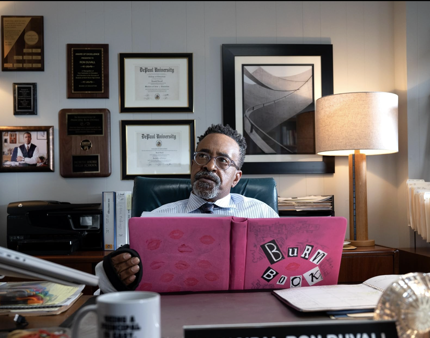 Principal Duvall (Tim Meadows) analyzing the 'Burn Book' given to him by Regina George (Reneé Rapp) in hopes of ruining Cady Heron's (Angourie Rice) reputation.
